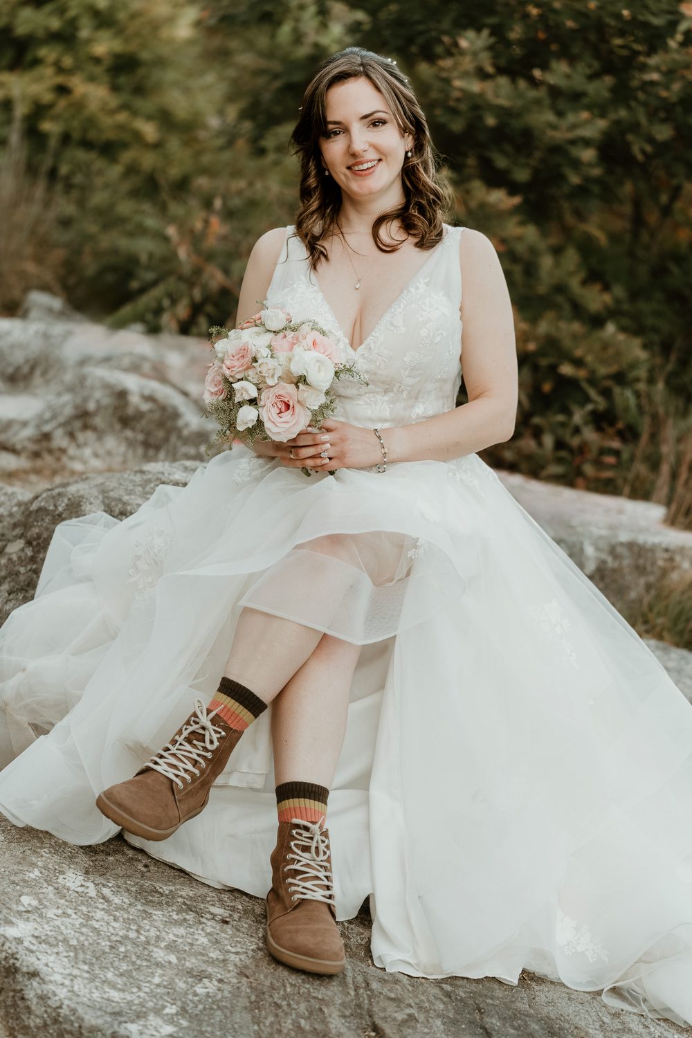 Bride in a gown and hiking boots holding a bouquet on a granite ledge in New England