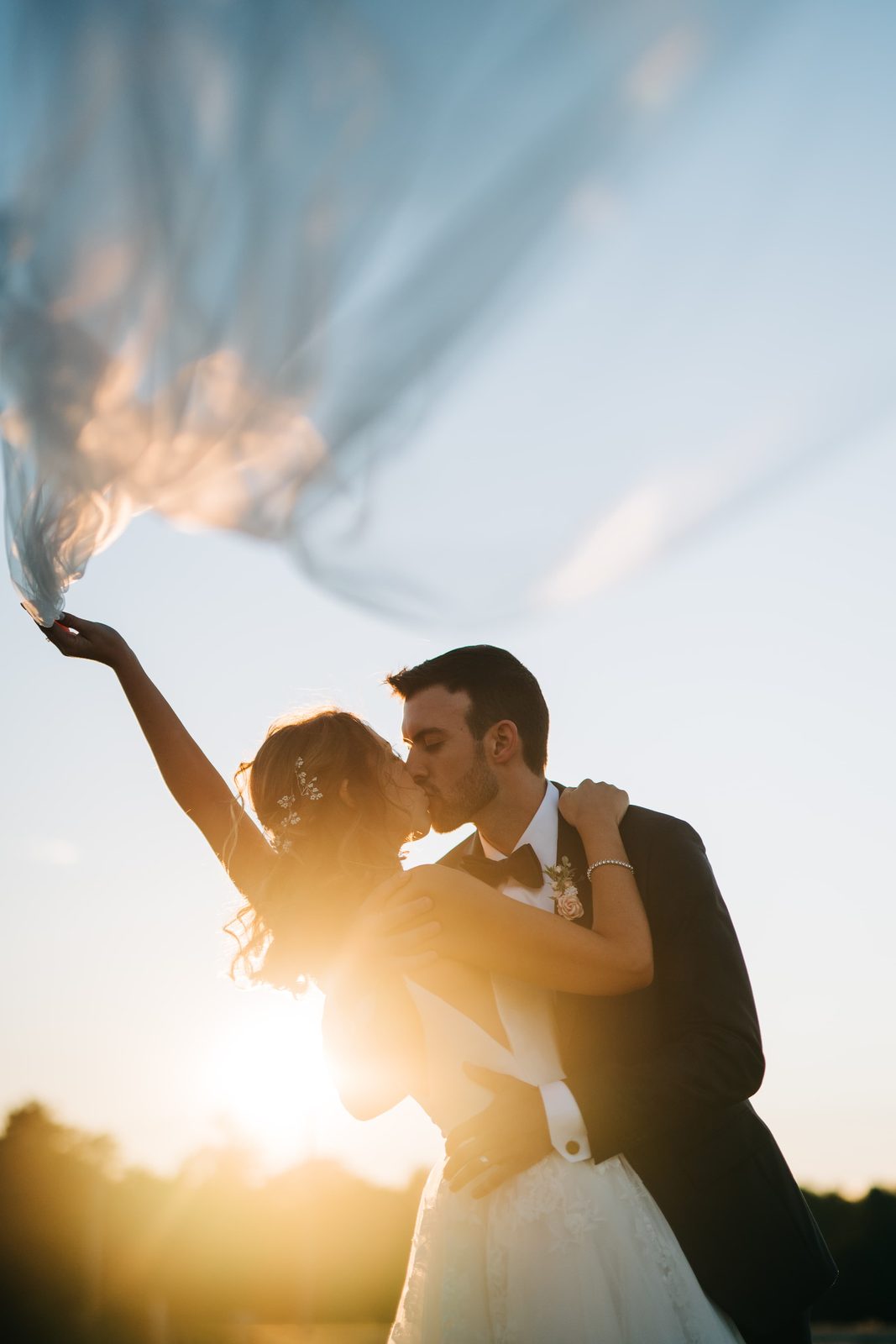 Bride and groom kissing at sunset as the veil sweeps into the sky